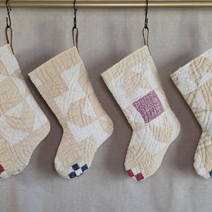 May include: Four quilted Christmas stockings hanging from a gold rod. Each stocking features a cream and white geometric pattern with intricate stitching. The toes of the stockings are accented with small, colorful checkered patches in red and blue.