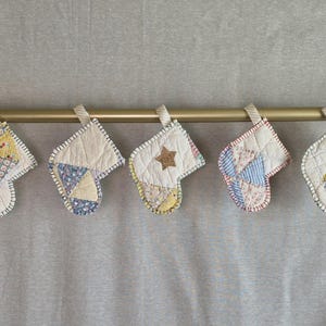 May include: Five small quilted stockings hanging from a gold rod. Each stocking features a unique patchwork design with various floral and geometric patterns in shades of yellow, blue, and white, finished with a decorative blanket stitch edge.