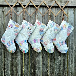 May include: Six quilted Christmas stockings with patchwork details in various colors, hanging from a rusty metal rack against a weathered wooden fence. The stockings have a cream-colored background and colorful patchwork accents.