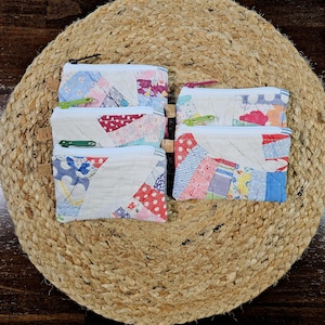 May include: A collection of five small, handmade patchwork zipper pouches. Each pouch features a unique design with a variety of colorful fabric scraps, including floral, polka dot, and striped patterns. The pouches have white zippers and are arranged on a woven, circular mat.