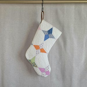 May include: A quilted Christmas stocking featuring a white background with a repeating star pattern in blue, orange, light green, and lavender floral prints. The stocking is suspended from a dark brown loop attached to a gold-toned metal rod.