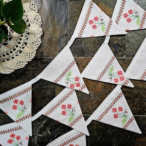 May include: White fabric bunting with red floral embroidery and green leaves. The bunting has a black and orange cross-stitch border. The design features a series of triangular flags connected by a white ribbon, perfect for festive decorations.