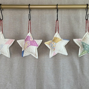 May include: Four star-shaped ornaments made from quilted fabric patches. Each star features a unique combination of white and colored fabric pieces, including blue, pink, yellow, and green patterns. They hang from black metal hooks on a rod.
