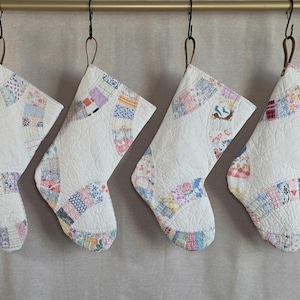 May include: Four quilted Christmas stockings hanging from a metal rod. Each stocking features a white base with curved, multi-colored patchwork fabric sections in various floral, plaid, and geometric patterns, finished with a stitched texture.