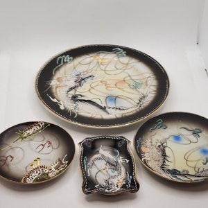 May include: Set of four black and white ceramic plates with gold trim and a dragon design. The plates are in various sizes and shapes, including a round plate, a square plate, and two smaller round plates.