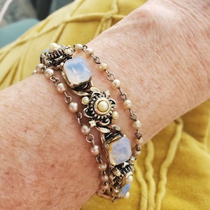 Op de afbeelding: Een zilveren armband met meerdere rijen nepparels en heldere glazen stenen. De armband heeft sierlijke metalen accenten met een bloemenmotief.