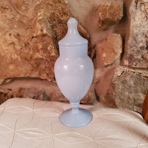 Könnte beinhalten: Eine hellblaue Glasdose mit Deckel und einem Sockel. Die Dose steht auf einer weißen Stoffoberfläche mit einem geometrischen Muster.