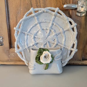 May include: A light blue ceramic wall pocket with a decorative plate and a small vase. The plate has a woven design, and the vase is adorned with a white rose and green leaves. The piece is mounted on a wooden door.