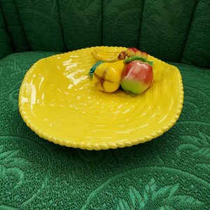 May include: A yellow ceramic fruit bowl with a raised, woven design. The bowl has a pear, apple, and banana sculpted on the rim.
