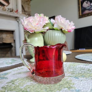 May include: A small, ruby red glass creamer with a clear handle and a textured base. The creamer has a ruffled rim and sits on a floral patterned placemat. In the background are two vases with pink flowers.