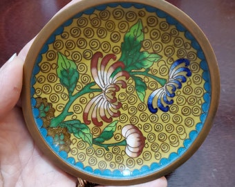 May include: A small, round, brass trinket dish with a blue and white floral design on a yellow background. The dish has a blue and white border.