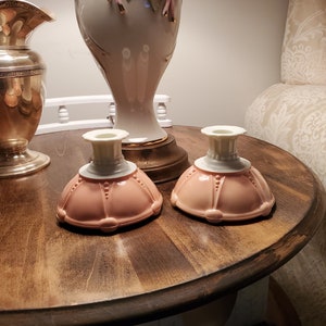 Peut inclure: Deux bougeoirs en céramique rose avec des dessus blancs et des accents dorés. Les bougeoirs sont sur une table en bois.