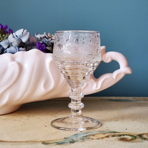 Puede incluir: Un vaso de chupito de vidrio transparente con un delicado patrón floral grabado en la superficie. El vaso tiene un tallo alto y delgado y una base redonda.