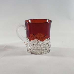 Puede incluir: Una taza pequeña de vidrio transparente con un interior rojo rubí. La taza tiene un asa de vidrio transparente y un diseño decorativo de vidrio cortado alrededor del borde.