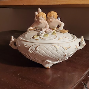 May include: A white porcelain sugar bowl with a lid featuring two cherubs holding flowers. The bowl has gold accents and a floral design.