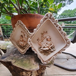 Puede incluir: Dos placas de pared decorativas con marcos ornamentados. Cada placa presenta una imagen en relieve de un niño en un entorno boscoso. Una placa representa a una niña con un vestido blanco y la otra a un niño con una chaqueta azul.