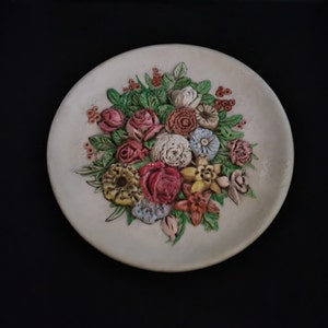 May include: A white ceramic plate with a raised floral design. The flowers are in shades of pink, red, yellow, and blue. The plate is a decorative piece and would be a great addition to any home.