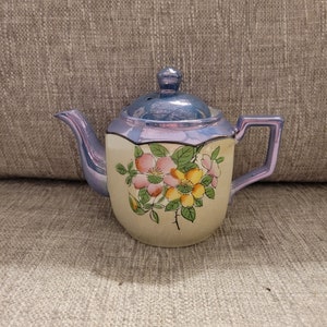 May include: A small, blue and white teapot with a floral design. The teapot has a lid and a handle. The flowers are pink, yellow, and green.