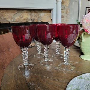 Puede incluir: Seis copas de vino tinto rubí con tallos transparentes y retorcidos sobre una mesa de madera. Las copas tienen una forma de cáliz clásica y están llenas de un líquido oscuro. Los tallos tienen un diseño decorativo y texturizado.
