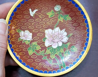 May include: A small, round, decorative dish with a red background and gold trim. The dish features a floral design with pink flowers, green leaves, and a white butterfly. The background is decorated with a repeating pattern of gold clouds.