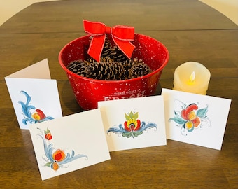 Puede incluir: Cuatro tarjetas de felicitación en blanco con diseños florales pintados a mano en rojo, amarillo, azul y verde. Las tarjetas están sobre una mesa de madera con un cuenco rojo lleno de piñas y una vela blanca.
