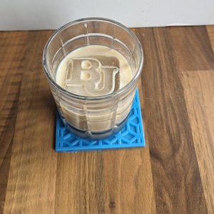 May include: A glass of light brown liquid with a large ice cube shaped like the letters "BJ" in the center. The glass is sitting on a blue silicone coaster.