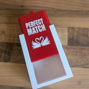 Könnte beinhalten: Eine rote Schachtel mit den Worten "PERFECT MATCH" in weißen Buchstaben. Unter dem Text befinden sich zwei weiße Schwan-Illustrationen. Die Schachtel befindet sich in einem weißen Rahmen, und der Hintergrund ist eine Holzoberfläche.