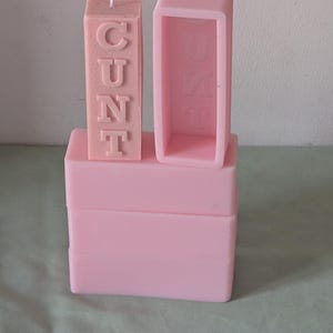 May include: Pink silicone candle mould with the word "CUNT" spelled out in raised letters. The mould is sitting on top of three other pink silicone moulds.