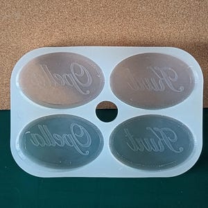 May include: A white silicone mold with four oval-shaped cavities. Each cavity has the word "Kurt" or "Opella" inscribed in a cursive font.