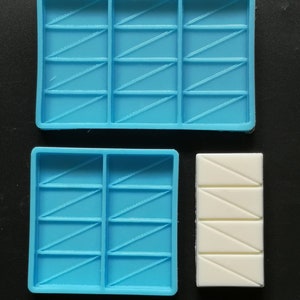 May include: Two blue silicone moulds for making chocolate bars. The moulds have a pattern of diagonal lines. One mould is larger than the other. A white chocolate bar is shown in the smaller mould.