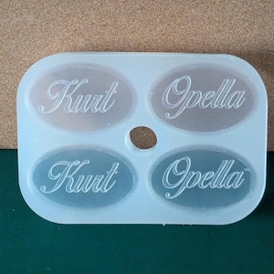 May include: A white silicone mold with four oval-shaped cavities. The mold is designed to create custom name tags or labels. The names "Kurt" and "Opella" are engraved into the mold.
