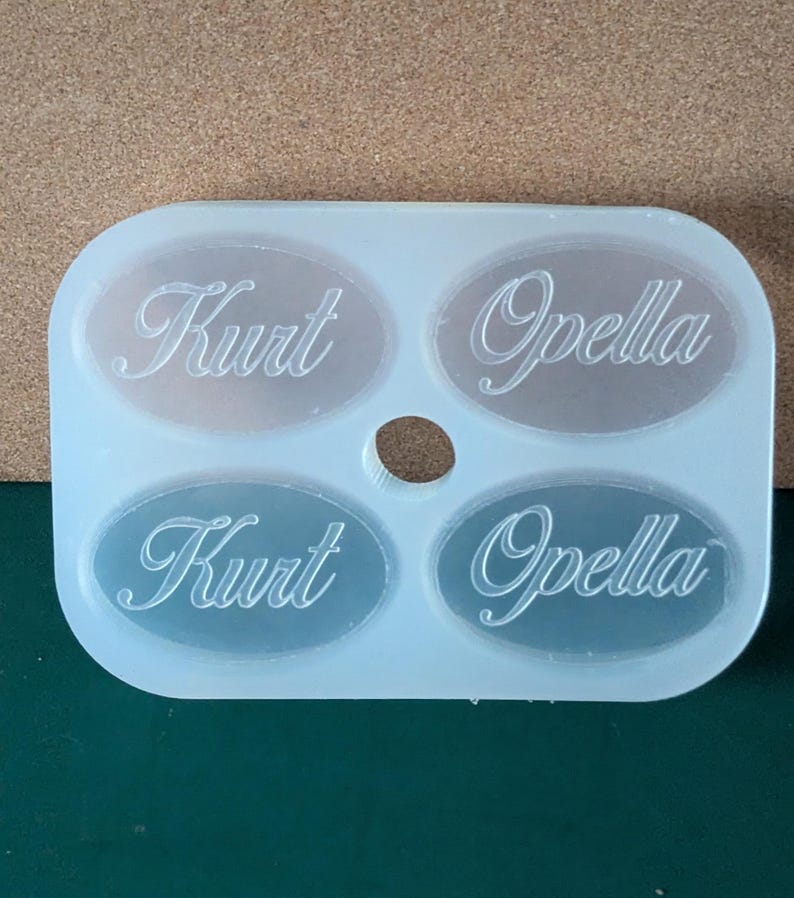 May include: A white silicone mold with four oval-shaped cavities. The mold is designed to create custom name tags or labels. The names "Kurt" and "Opella" are engraved into the mold.