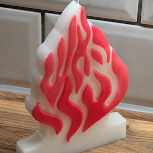 Peut inclure: Une bougie blanche en forme de flamme avec des accents de cire rouge. La bougie a une base plate.