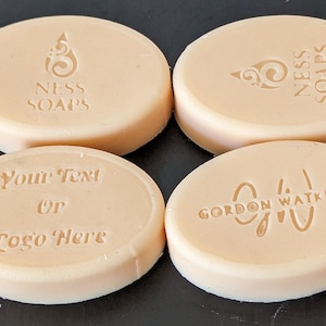May include: Four oval-shaped bars of soap with different designs. The first bar has the text "NESS SOAPS" and a swirl design. The second bar has the text "NESS SOAPS" and a swirl design. The third bar has the text "Your Text or Logo Here". The fourth bar has the text "GORDON WATKINS" and a swirl design.