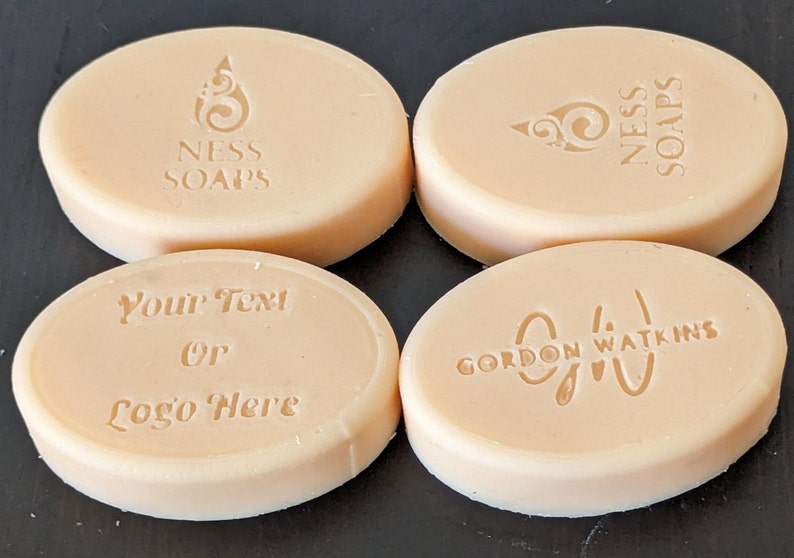 May include: Four oval-shaped bars of soap with different designs. The first bar has the text "NESS SOAPS" and a swirl design. The second bar has the text "NESS SOAPS" and a swirl design. The third bar has the text "Your Text or Logo Here". The fourth bar has the text "GORDON WATKINS" and a swirl design.