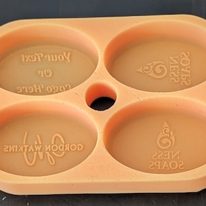 May include: Orange silicone soap mold with four oval-shaped cavities. The mold has a hole in the center. The text "SOAPS NESS" is printed in the top right cavity. The text "SOAPS NESS" is printed in the bottom right cavity. The text "GORDON WATKINS" is printed in the bottom left cavity. The text "LOGO Here? Or Your Text" is printed in the top left cavity.