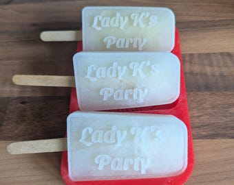 May include: Three translucent, off-white popsicle-shaped items with wooden sticks, each featuring the white text Lady K's Party. These items are arranged horizontally and nested within a bright red, rectangular silicone mold tray.