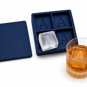 May include: A navy blue silicone ice cube tray with the letter "A" imprinted on each cube. One ice cube is shown outside the tray, and another is in a rocks glass filled with amber liquid. A matching navy blue lid is also present.