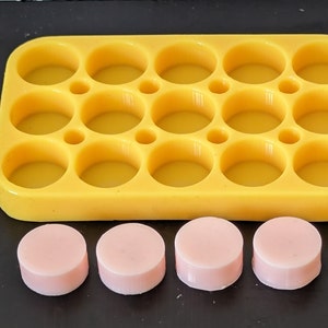 May include: A yellow silicone mould with 12 round cavities, and four pink round soap bars sitting on a black surface.