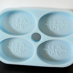 May include: A light blue silicone mold with four oval-shaped cavities. Each cavity has the text "Gordon Watkins" embossed in the bottom.
