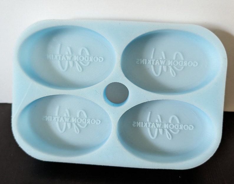May include: A light blue silicone mold with four oval-shaped cavities. Each cavity has the text "Gordon Watkins" embossed in the bottom.