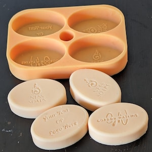 Peut inclure: Quatre barres de savon ovales dans un moule en silicone. Les barres de savon sont blanches et présentent différents textes et motifs. Le texte sur les barres de savon comprend "Your Text or Logo Here", "Gordon Watkins" et "Niss Soaps".