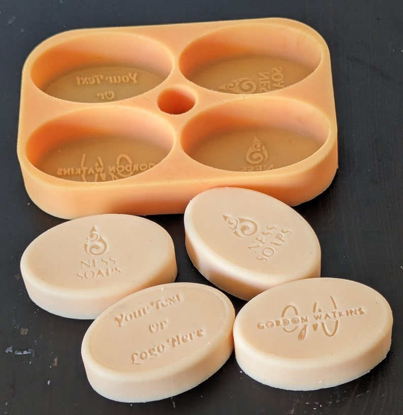 May include: Four oval-shaped soap bars in a silicone mold. The soap bars are white and have different text and designs. The text on the soap bars includes "Your Text or Logo Here", "Gordon Watkins", and "Niss Soaps".