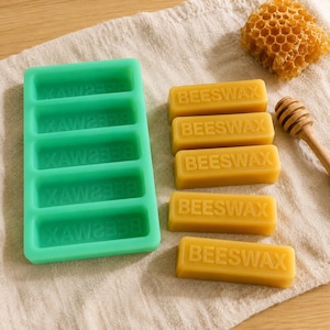 Può includere: Uno stampo rettangolare in silicone verde menta con cinque scomparti, cinque barrette rettangolari di cera d'api gialla con la scritta BEESWAX in rilievo, un pezzo di favo dorato e un bastoncino per miele in legno su un tessuto.