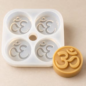 May include: A square, translucent silicone mold featuring four circular cavities, each embossed with the Om symbol. Beside the mold sits a single, circular, tan-colored solid piece cast from the mold, displaying a raised Om symbol on its surface.