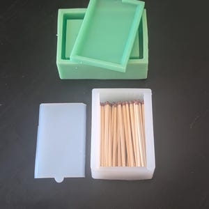 Op de afbeelding: Een set van drie siliconen mallen in groen- en wittinten. Eén groene mal is rechthoekig met een deksel, een andere is wit en rechthoekig, en de derde is wit en rechthoekig en gevuld met houten lucifers.