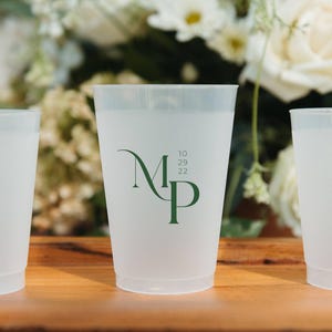 May include: A set of three translucent frosted plastic cups featuring a dark green printed monogram with the letters M and P, accompanied by the date 10 29 22. The cups have a tapered cylindrical shape with a smooth rim and a flat base.