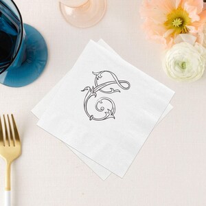May include: A square white paper napkin featuring a centered black decorative letter G in a swirling calligraphic script. The napkin has a textured border and is positioned near a gold-toned fork and a blue glass base on a light surface.