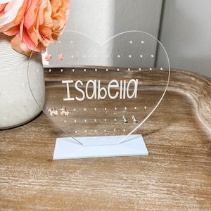 May include: A clear acrylic heart-shaped earring holder with a white base. The holder has multiple holes for earrings and the name "Isabella" is printed on the front.