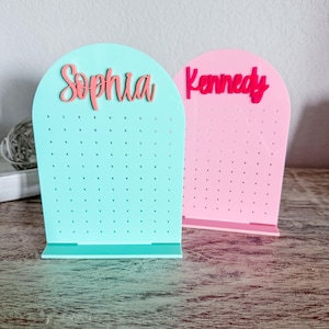 May include: Two earring display stands, one aqua and one pink, each with a name in a contrasting color. The aqua stand has the name "Sophia" in pink, and the pink stand has the name "Kennedy" in red. Both stands have multiple small holes.